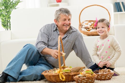 Grandfather and granddaughter sitting on a carpet in the living room next to woven baskets with pastries.