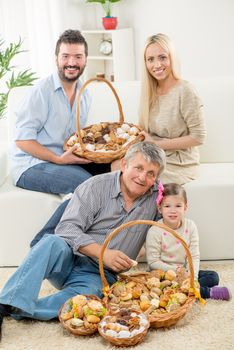 Senior man and a little girl are sitting on the living room floor. In front of them is basket with pastries, and behind them young parents are sitting on the couch. Looking at camera.