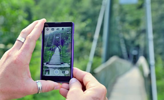 View over the mobile phone display during taking a picture of bridge in nature. Man is holding the mobile phone in hand and taking a photo in nature. Closeup view on hands with mobile phone.