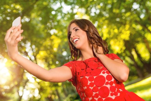 Young beautiful woman taking a selfie in red dress and enjoy the sun in the park. 