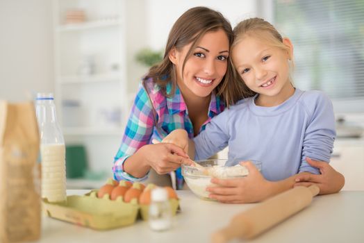 Beautiful mother and her daughter making dough together in the domestic kitchen. Looking at camera.