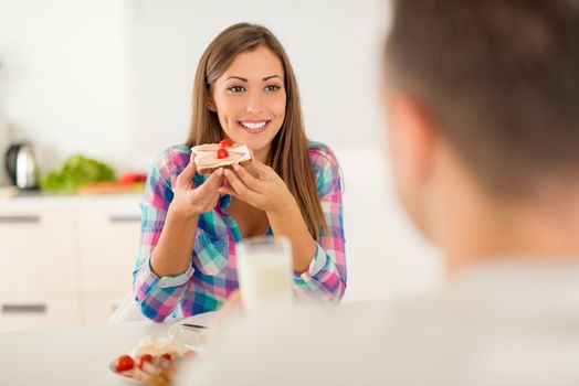 Beautiful young couple having breakfast in the domestic kitchen. Girlfriend eating sandwich and looking her boyfriend.