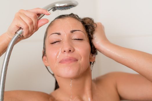 Beautiful young smiling woman washing face and hair while showering under shower head.