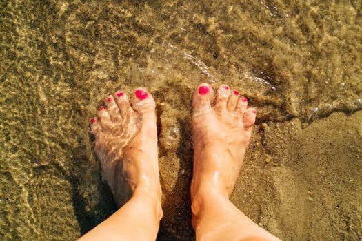 Woman standing in the coast on the sandy beach. Close-up. Focus on a foot. Top view.