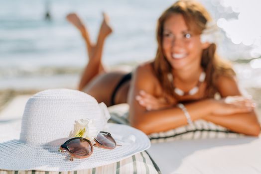Beautiful young woman enjoying on the beach. She is smiling and looking away. In foreground is white summer hat and sunglasses. Selective focus. Focus on foreground.