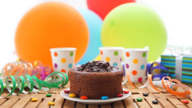 Chocolate birthday cake on rustic wooden table with background of colorful balloons, gifts, plastic cups with candies and white wall in the background