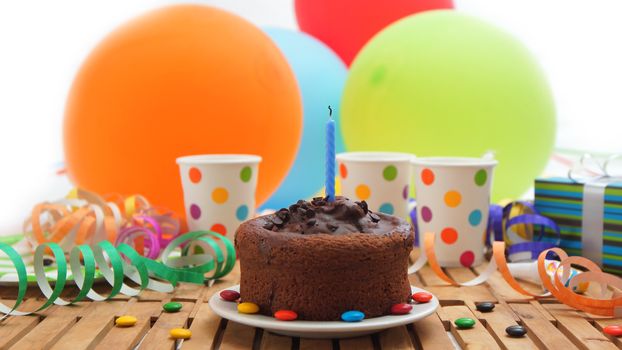 Chocolate birthday cake with a blue candle extinguished on rustic wooden table with background of colorful balloons, gifts, plastic cups with candies and white wall in the background