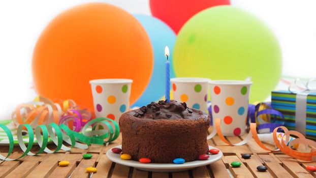 Chocolate birthday cake with a blue candle burning on rustic wooden table with background of colorful balloons, gifts, plastic cups with candies and white wall in the background