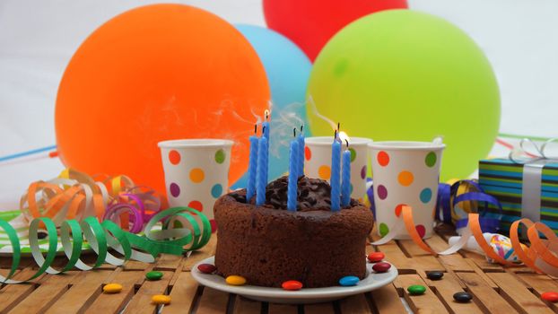 Chocolate birthday cake with a blue candles extinguished on rustic wooden table with background of colorful balloons, gifts, plastic cups with candies and white wall in the background