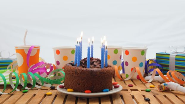 Chocolate birthday cake with a blue candles burning on rustic wooden table with background of colorful streamers, gifts, plastic cups with candies and white wall in the background