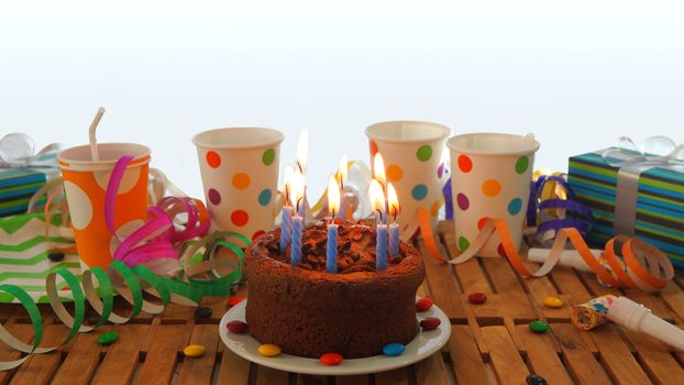 Chocolate birthday cake with a blue candles burning on rustic wooden table with background of colorful streamers, gifts, plastic cups with candies and white wall in the background