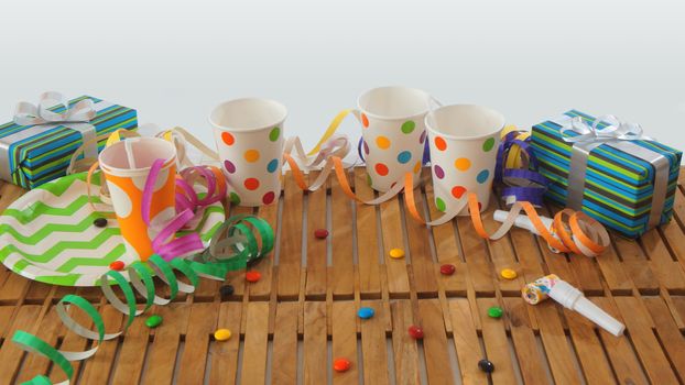 Rustic wooden table with colorful streamers, gifts, plastic cups, plastic plate, candies on white background. Party background