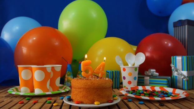 Birthday cake on rustic wooden table with background of colorful balloons, gifts, plastic cups and plastic plate with candies and blue wall in the background