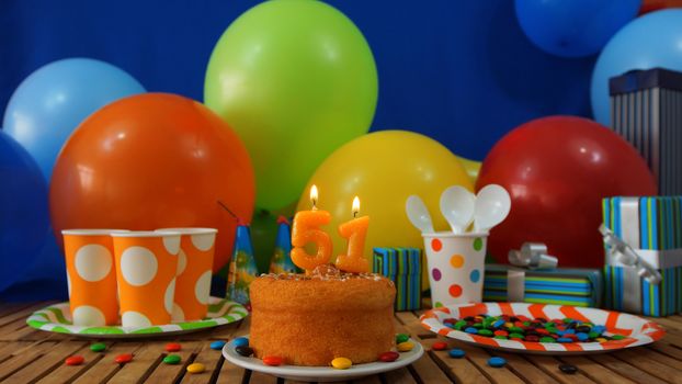 Birthday cake on rustic wooden table with background of colorful balloons, gifts, plastic cups and plastic plate with candies and blue wall in the background