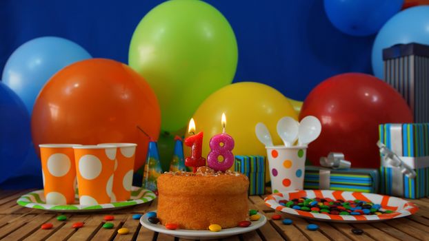 Birthday cake on rustic wooden table with background of colorful balloons, gifts, plastic cups and plastic plate with candies and blue wall in the background