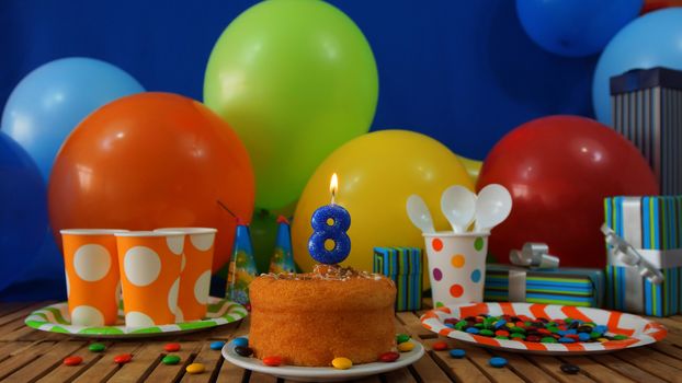 Birthday cake on rustic wooden table with background of colorful balloons, gifts, plastic cups and plastic plate with candies and blue wall in the background