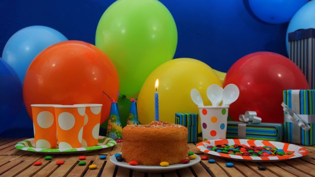 Birthday cake with a blue candle burning on rustic wooden table with background of colorful balloons, gifts, plastic cups and plastic plate with candies and blue wall in the background