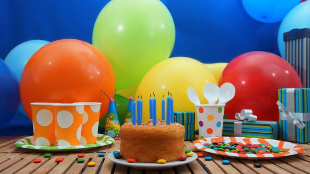 Birthday cake with a blue candles extinguished on rustic wooden table with background of colorful balloons, gifts, plastic cups and plastic plate with candies and blue wall in the background