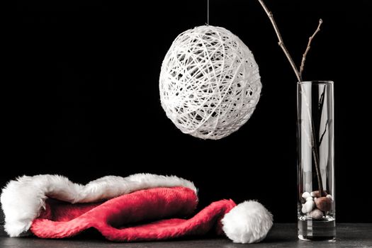 Alder branch in the glass with a white christmas ball and santa claus hat on the black stone table horizontal