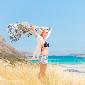 Relaxed woman, arms rised, holding colorful scarf, enjoying sun, freedom and life at beautiful beach. Young lady feeling free, relaxed and happy. Concept of vacations, freedom, joy and well being.