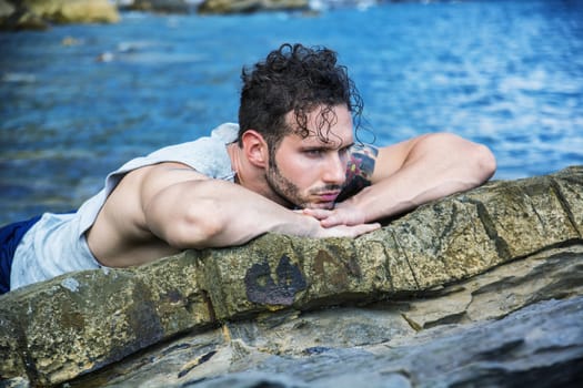 Handsome muscular man on the beach lying on rocks, looking at camera