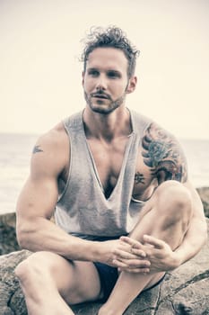 Handsome muscular man on the beach sitting on rocks, looking at camera