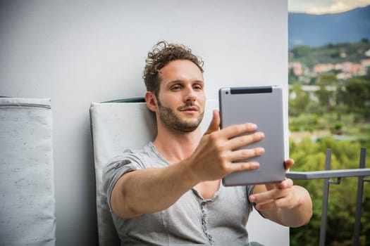 Handsome trendy man wearing t-shirt sitting and doing videochat, looking down at a tablet computer that he is holding, outdoor on house terrace