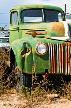 An old truck sitting in a junkyard with weeds growing up around it.