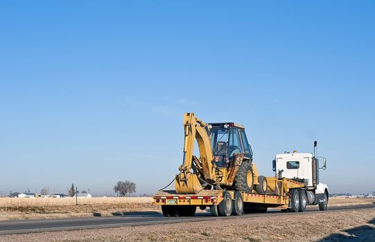 Big truck with a drop-deck trailer hauling a back-hoe tractor.