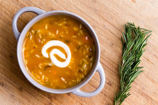 Cup of chicken broth with wild rice served with a twirl of sour cream ready to be garnished with fresh rosemary displayed alongside on a bamboo cutting board, overhead view