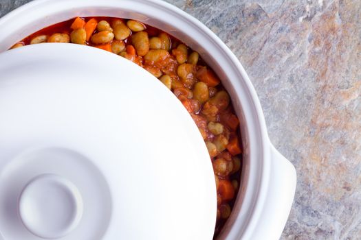 Delicious savory lima bean casserole with carrots cooling off in a white ceramic casserole dish with the lid moved aside to view the contents, closeup overhead view on a stone counter