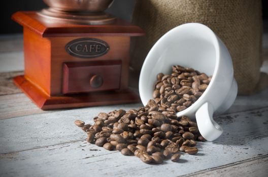 Coffee grain spill from a cup. Jute bag of roasted beans and mill in background