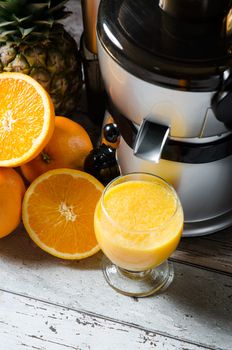 Juicer and orange juice in glass on wooden desk. Fruits in background