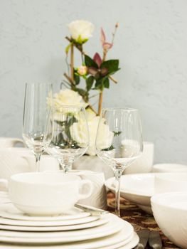 Set of new dishes on table with tablecloth. Stack of white plates and wine glasses with flowers on restaurant table. Vertical. Shallow DOF
