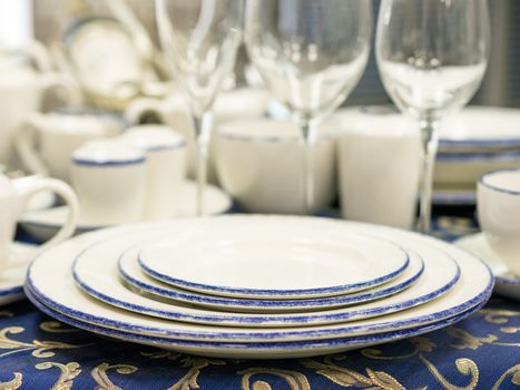Set of new dishes on table with blue tablecloth. Stack of plates, saucer and wine glasses on restaurant table. Shallow DOF
