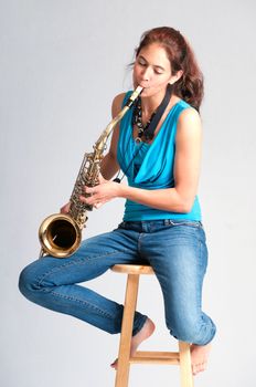 Pretty brunette girl practicing on the saxophone while seated barefooted on a stool