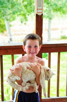 Cute little boy with no shirt in the summer carrying a cat in his arms.