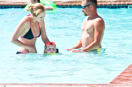 A young couple showing their little boy how to swim with a safety flotation device on.
