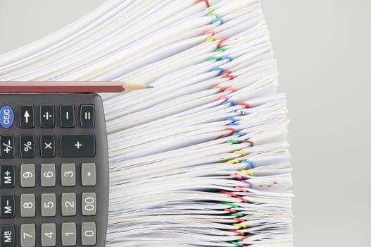 Brown pencil place on calculator in vertical and blur overload of paperwork with colorful paperclip with white background.