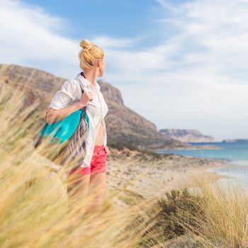 Relaxed woman on vacations in white loose shirt carrying beach bag and towel, enjoying beautiful coastline view of white sandy lagoon at Balos beach, Greece.