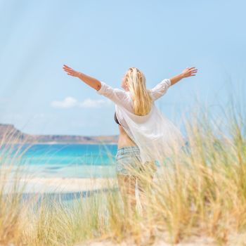 Relaxed woman, arms rised, enjoying sun, freedom and life an a beautiful beach. Young lady feeling free, relaxed and happy. Concept of vacations, freedom, happiness, enjoyment and well being.