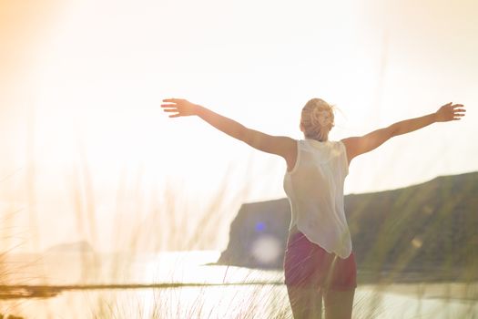 Relaxed woman in white shirt, arms raised, enjoying sun, freedom and life an beautiful beach. Young lady feeling free, relaxed and happy. Concept of vacations, freedom, happiness, joy and well being.