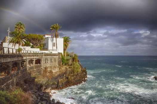 Town city hall over rocks and sea waves under a rainbow in Puerto de la Cruz, Tenerife, Spain