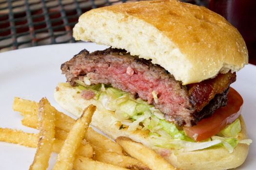 Grilled hamburger with bacon, shredded lettuce, tomatoe slice.  Meat on bread roll with fried potatoes on outdoor, wrought iron patio table.  