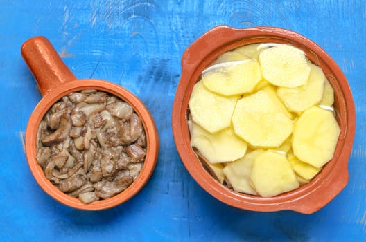 Sauteed mushrooms and raw sliced potatoes in the ceramic utensils for cooking. Blue wooden background.