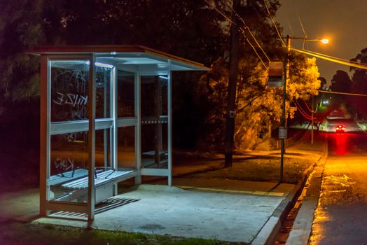 Empty bus shelter at night.  A car is dissappearing down the road.  graffiti is scratched into the glass.