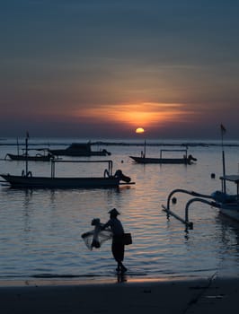 Man fishing with a net in Bali with traditional boats in the background.