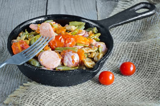 Fried vegetables with sliced sausage, in an old cast iron skillet. A piece of sausage on a fork and gray wooden background.