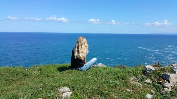 blonde girl sitting back on the rock, looking on the water