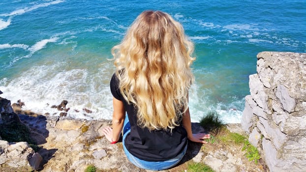 blonde girl sitting back on the rock, looking on the water